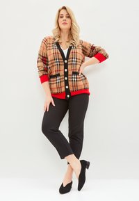 Beżowy kardigan w kratkę z czerwonymi mankietami i dołem, z dużymi guzikami i dekoltem w serek. Zestawiony z czarnymi spodniami slim fit i czarnymi butami.