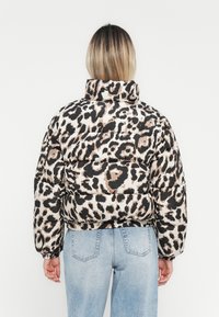 Pufferjack met een bruin en zwart luipaardprint, hoge kraag en elastische manchetten, gecombineerd met lichtblauwe jeans.