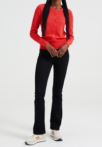 Cardigan rosso con texture e chiusura a bottoni, abbinato a jeans neri a zampa d'elefante e scarpe da ginnastica beige con dettagli arancioni.