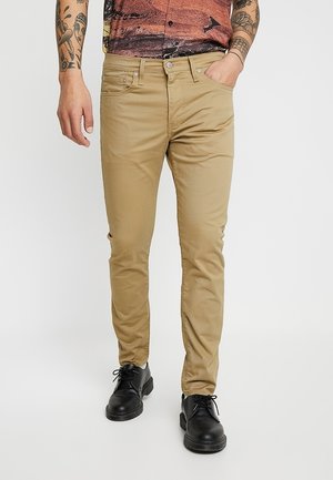 Mężczyzna w beżowych, dopasowanych spodniach, czarnych sznurowanych butach i wzorzystej koszuli z krótkim rękawem, z tatuażami na ramionach, stojący z jedną ręką w kieszeni.