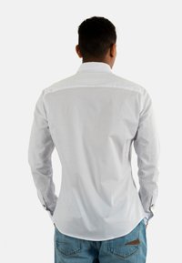 Chemise blanche à manches longues et à boutonnière, avec une texture lisse et un ourlet arrondi. Elle se caractérise par un col classique et des manchettes à boutons, portée avec un jean bleu.