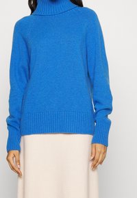 Donna che indossa un maglione a collo alto a coste blu brillante con maniche lunghe e una gonna beige chiaro su uno sfondo neutro.