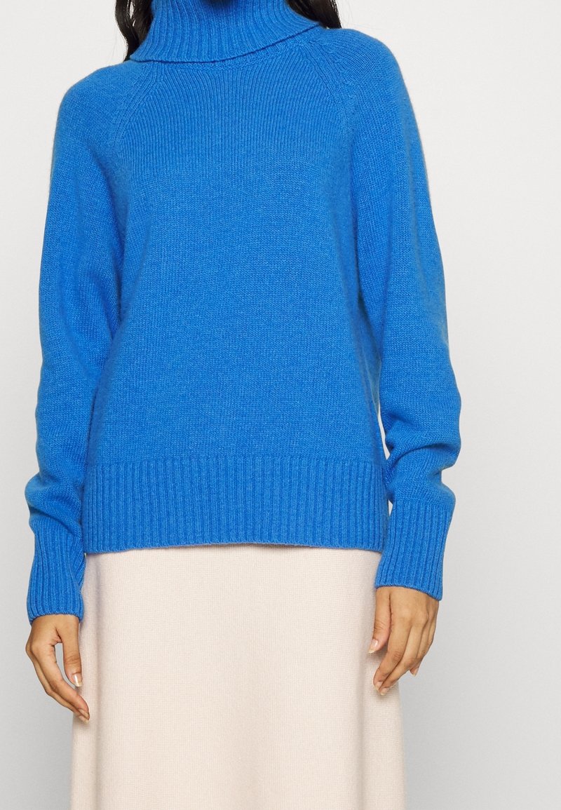 Donna che indossa un maglione a collo alto a coste blu brillante con maniche lunghe e una gonna beige chiaro su uno sfondo neutro.