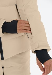 Beige puffjacka med dragkedja och kardborrefäste vid ärmarna. En svart figurnära ärm syns under jackans arm. Slät textur.