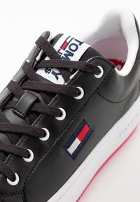 Černé kožené tenisky s hnědými tkaničkami, bílými očky a bílým jazykem s logem. Na boku je barevná emblema a bílá podrážka.