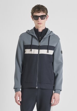 Joven con gafas de sol negras y una sudadera azul con cremallera que tiene rayas horizontales en blanco y negro, de pie contra un fondo claro y liso.