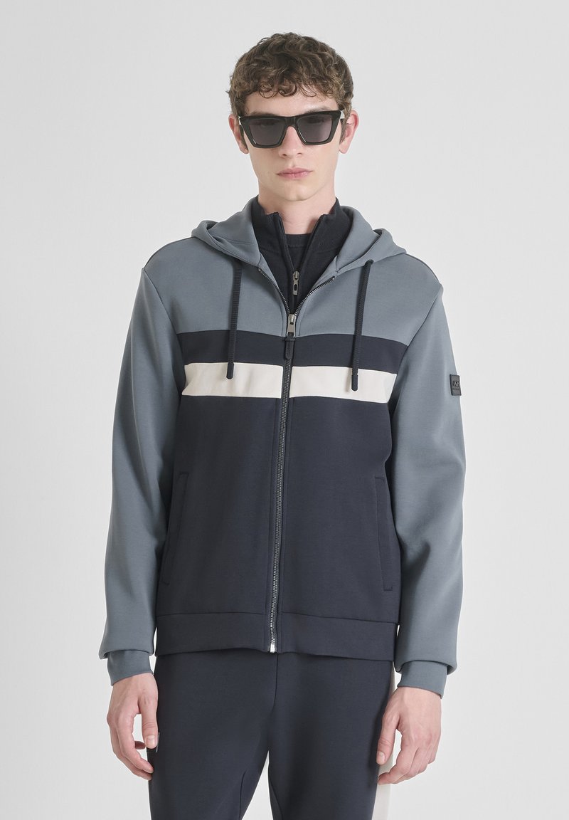 Giovane vestito con occhiali da sole neri e una felpa blu con zip a righe orizzontali bianche e nere, in piedi contro uno sfondo chiaro e uniforme.
