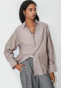 Svetlohnedá a krémová pruhovaná ko šeľa na gombíky s voľným strihom a dlhými rukávmi, kombinovaná so sivými nohavicami. Má špicatý golier a bočnú kapsu.