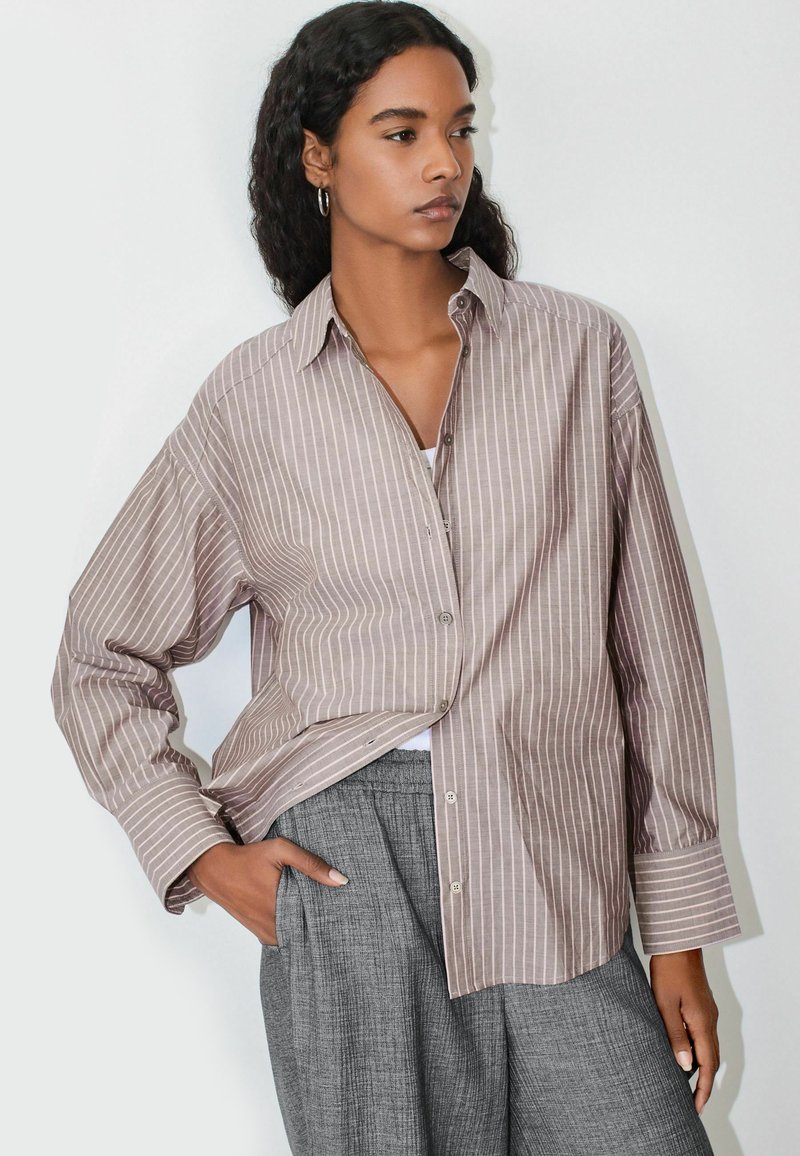 Camisa abotonada de rayas en marrón claro y crema con un ajuste relajado y mangas largas, combinada con pantalones grises. Presenta un cuello puntiagudo y un bolsillo lateral.