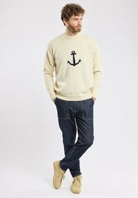 Pull crème avec un motif d'ancre noire, associé à un pantalon en jean bleu foncé et des chaussures beiges. Le pull a un col et des poignets à côtes.