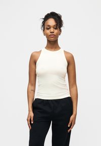 Jeune femme aux cheveux bouclés portant un haut blanc sans manches et un pantalon noir, debout devant un fond clair uni.