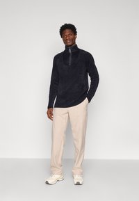 Svart fleece-pullover med halvvinddrag, kombinerad med ljusa beige cargobyxor och vita sneakers. Enkel, mjuk textur och modern design.