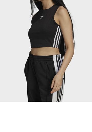 Vrouw draagt zwarte Adidas crop top en zwarte trainingsbroek met witte zijstrepen en drukknopen, staand tegen een effen achtergrond.