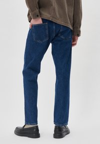 Vaqueros de mezclilla azul con un corte recto, que presentan un diseño de bolsillo trasero y sutilezas en los detalles de costura, combinados con unos zapatos negros de tipo slip-on.