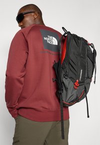 Sort og grå rygsæk med røde detaljer, buet form, flere rum og justerbare stropper, kombineret med en bordeaux sweatshirt.
