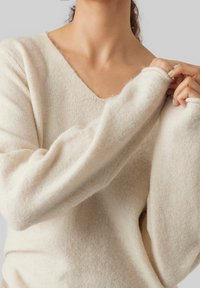 Beżowy sweter na drutach z dekoltem w serek, długimi rękawami, miękką fakturą i lekko luźnym krojem. Posiada ściągacze przy mankietach.