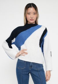 Chunky strikkegenser med et geometrisk mønster i marineblått, blått og hvitt. Designet med høy hals og vide ermer.