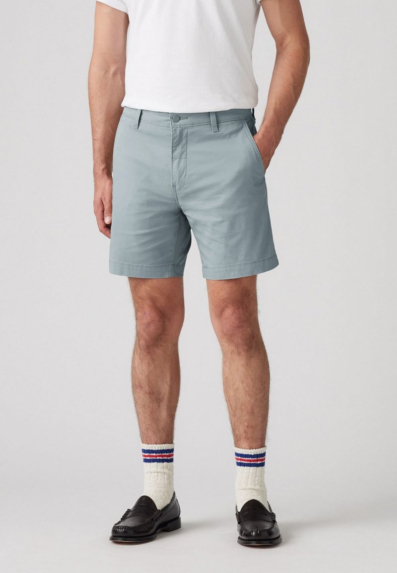 Shorts bleu clair en tissu lisse, dotés d'une fermeture à bouton et à glissière, associés à des mocassins noirs et des chaussettes blanches à rayures colorées.