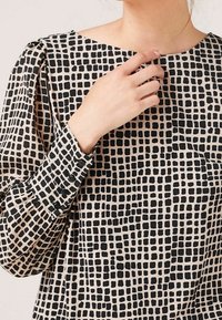 Schwarze und beige Bluse mit kariertem Muster, weichem Stoff, rundem Ausschnitt und geknöpften Manschettendetails an den langen Ärmeln.