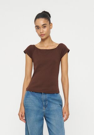 Kvinde med håret i en knold iført en tætsiddende kortærmet brun top og løstsiddende blå jeans, stående mod en ensfarvet lys baggrund.