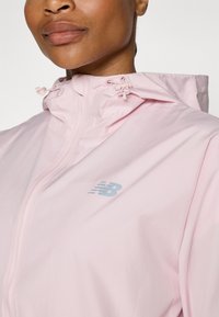Light pink windbreaker jacket with a hood, featuring a zippered front and a small blue New Balance logo on the chest. Smooth texture.