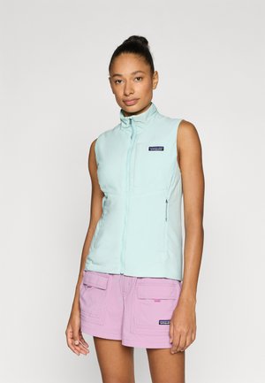 Veste sans manches vert menthe clair avec un col montant, une fermeture éclair à l'avant, des poches latérales et un écusson logo bleu marine. Portée avec un short rose pâle.