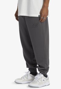 Man wearing dark gray DC sweatpants, white DC sneakers, black socks, and a white shirt, with rings on fingers visible.