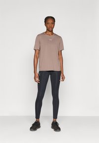 Καφέ κοντομάνικο μπλουζάκι με στρογγυλή λαιμόκοψη και μικρό λογότυπο στο στήθος, συνδυασμένο με μαύρο κολάν και μαύρα αθλητικά παπούτσια.