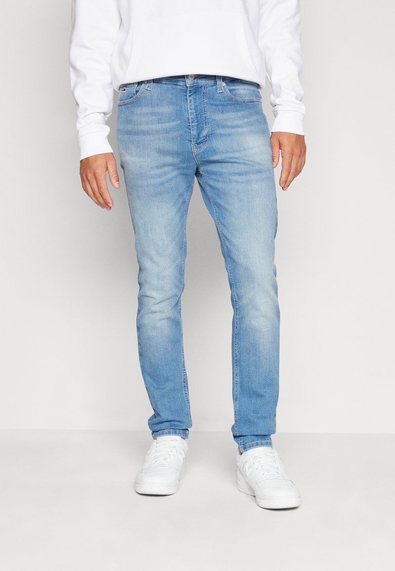 Világoskék, slim fit farmernadrág finom kopással. Denim anyagból készült, öt zsebbel és cipzáros záródással, fehér sportcipőkkel párosítva.