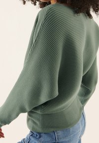 Grüner, gerippter Pullover mit überschnittenen Schultern und einem figurbetonten, gerippten Saum. Weiche Textur, leicht und lässig, kombiniert mit hoch taillierten Jeans.