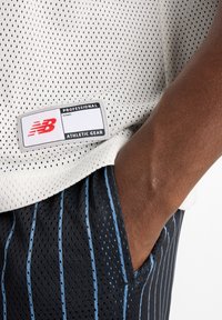 White mesh athletic shirt with black dotted pattern, featuring a grey tag. Black shorts with vertical blue stripes and a side pocket.