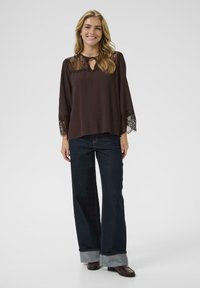 Brown blouse with lace accents on sleeves, round neckline with keyhole opening. Paired with dark wide-leg jeans featuring rolled cuffs.