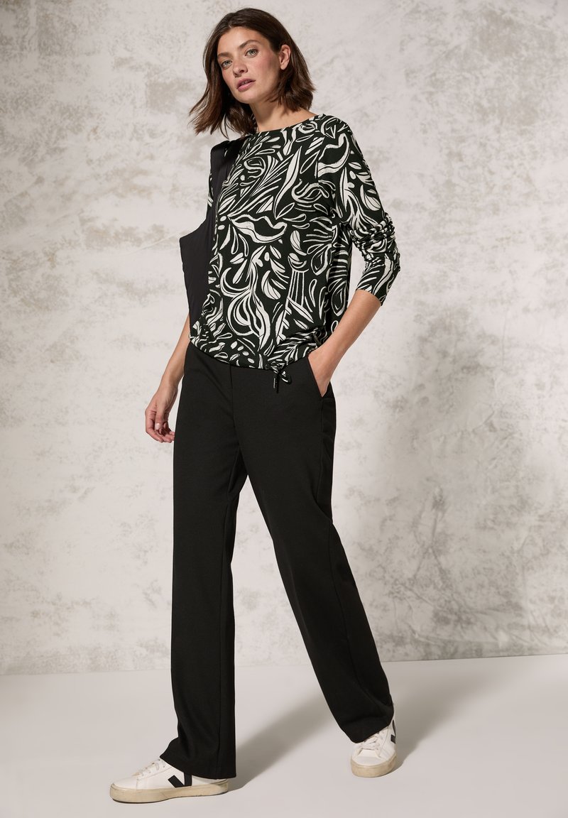 Langärmliges Oberteil mit schwarz-weißem Muster, kombiniert mit schwarzen Hosen und weißen Sneakern. Glatter Stoff mit lässiger Passform und seitlichen Taschen.