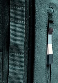 Tmavě zelený látkový batoh s texturovanými popruhy, se zipovou kapsou a barevným zatahovacím táhlem. Materiál vykazuje vodní kapky.