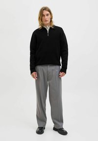 Mladý člověk s dlouhými blond vlasy má na sobě černý svetr s krátkým zipem, šedé skládací kalhoty a černé boty, stojí proti jednoduchému bílému pozadí.