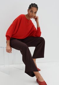 Pull rouge surdimensionné avec épaules tombantes, associé à un pantalon large marron et des chaussures rouges à enfiler. Chaise en acrylique transparente en arrière-plan.