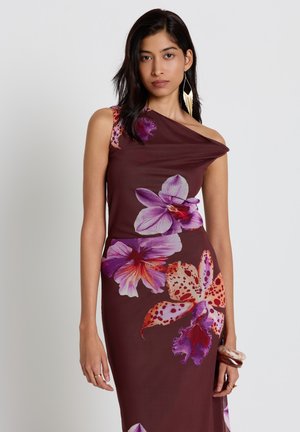 Femme portant une robe bordeaux à une épaule avec de grands imprimés floraux roses et violets, accessoirisée de boucles d'oreilles pendantes et de bracelets.