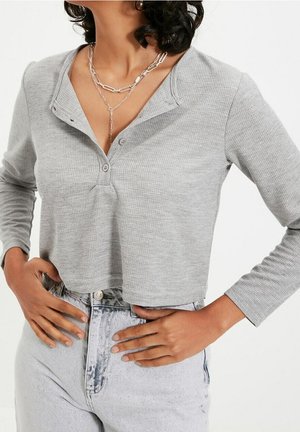 Gray cropped long-sleeve henley shirt with textured fabric, three buttons at the collar, and a relaxed fit, paired with light denim jeans.