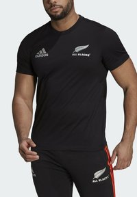 Musta lyhythihainen t-paita, jossa on Adidas-logo ja "All Blacks" -symboli valkoisena. Valmistettu kevyestä, sileästä kankaasta.