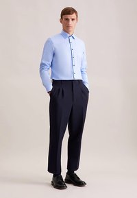 Lichtblauw overhemd met zwarte knopen, gecombineerd met donkerblauwe geplooide broek en zwarte schoenen, tegen een neutrale achtergrond.