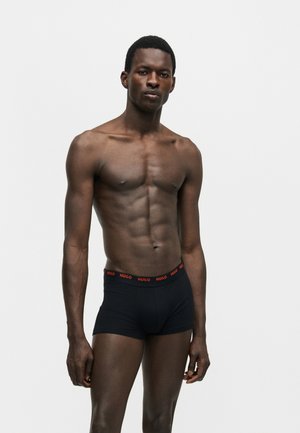 Svalnatý muž stojaci pred jednofarebným pozadím, nosiace čierne boxerky s červeným pásom s logom "HUGO".