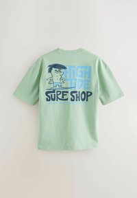 Lys grøn t-shirt med korte ærmer, prydet med en tegneserie grafik og teksten "HIGH TIDE SURF SHOP MANHATTAN BEACH" på bagsiden.