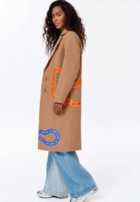 Žena s kudrnatými vlasy má na sobě béžový kabát s oranžovými a modrými nápisy "NEZASTAVUJ", široké světle modré džíny a stříbrné tenisky, stojí bokem.