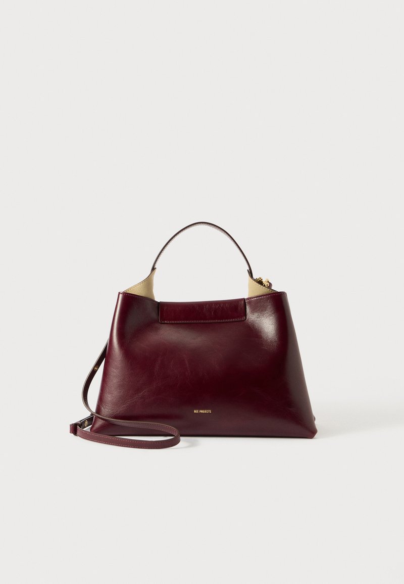 Bordeaux læder håndtaske med en struktureret form, kort håndtag og en aftagelig rem. Har et guld-logo foran og en glat tekstur.