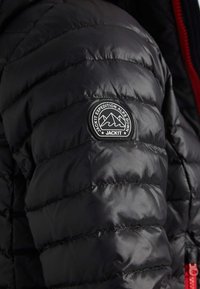 Sort pufferjakke med en kviltet overflade, et rundt logo-patch på ærmet og røde detaljer langs lynlåsen.