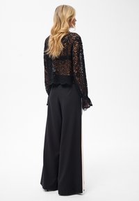 Blondynka ubrana w czarn�ą koronkową bluzkę z długim rękawem oraz czarne szerokie spodnie z białymi paskami po bokach, stojąca na białym tle.