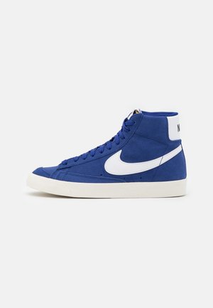 Zapatilla alta azul con suela blanca y logo Nike swoosh blanco sobre un fondo claro y liso, vista lateral.