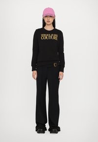 Melnā džemperī ar zelta "VERSACE JEANS COUTURE" tekstu, apvienots ar melniem platiem biksēm un rozā beisbola cepuri. Gluda auduma struktūra.
