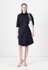 Navy blue, knee-length shirt dress with short sleeves, a belted waist, and button-down front. Model carries a small white handbag.