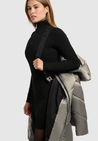 Schwarzes, geripptes Strickkleid mit hohem Kragen, kombiniert mit einem grauen, gesteppte Mantel, der über der Schulter getragen wird. Auffällige Metallbeschläge an den Mantelriemen.